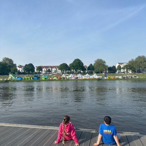 Zwei Personen auf einem Steg am Fluss sitzen. Eine Person trägt pink, die andere blau.
